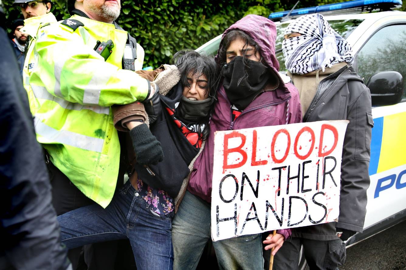 A protestor is arrested in front of a police car while another protestor holds a sign that reads "Blood on their hands".
