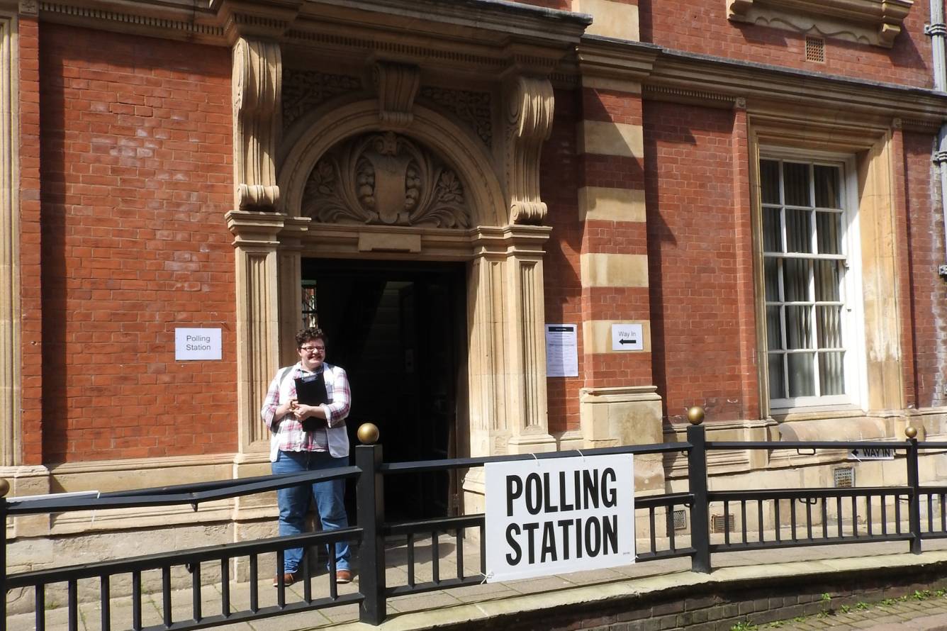 A polling station in Leicester city centre with a poll worker stood outside with a clipboard. They are smiling at the camera and there is a big sign on the railings in front of them that reads "Polling Station".